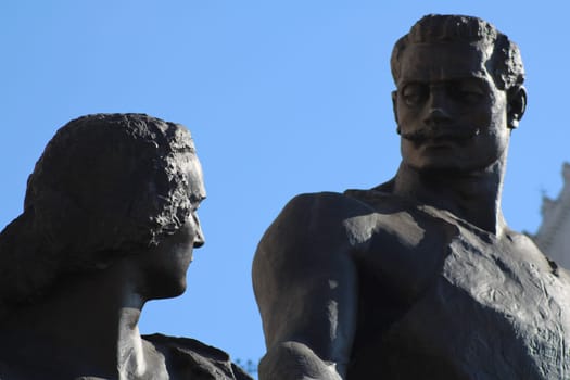 A striking statue depicts a man and woman locked in an intimate gaze, set against a bright blue sky in an urban environment. The artwork captures human connection and emotion.