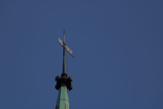 The towering spire of a church stands proudly against the bright blue sky, with a decorative weather vane twirling gently in the midday breeze, showcasing intricate design details.
