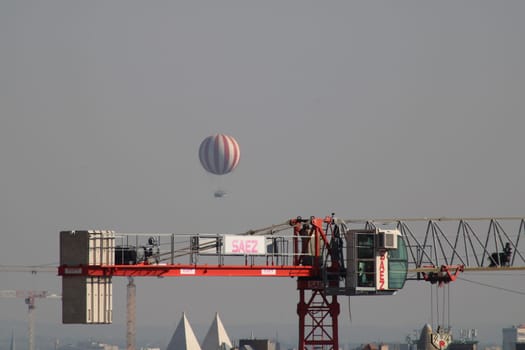A vibrant hot air balloon floats peacefully above a construction crane, creating a striking contrast against the clear blue sky. The urban landscape adds depth to the scene.