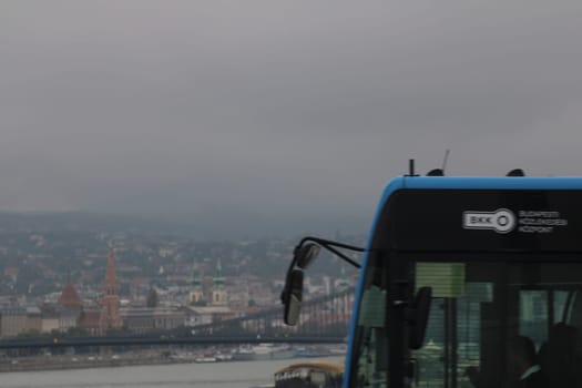 A blue bus is parked at a scenic viewpoint, providing a cloudy backdrop of Budapest and the Danube River. The cityscape features historic buildings and bridges.