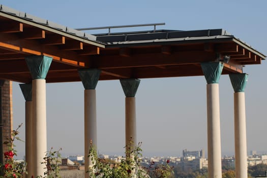 A beautifully designed wooden terrace features detailed columns and a clear view of a bustling cityscape under a bright blue sky during the day.