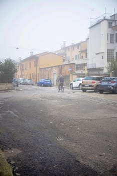 Cremona, Lombardy, Italy - November 7th 2024 Senior man riding a bicycle on a foggy street in a residential neighborhood with parked cars