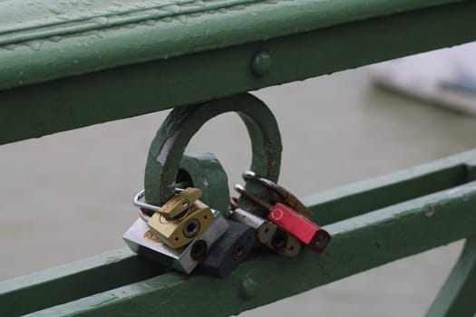 A variety of colorful love locks are secured to a green bridge railing, overlooking a river. The overcast weather adds a tranquil atmosphere to this moment.