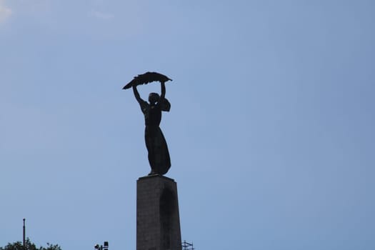 A tall statue depicting a figure holding a bird on a pedestal stands gracefully against a bright blue sky. This serene urban park captures the tranquil essence of the moment.