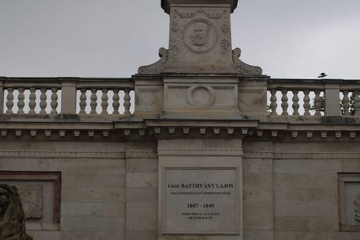 A detailed stone monument honors Batthyány Lajos, featuring ornate carvings and inscriptions. The surrounding area is calm with gray skies and lush greenery, inviting reflection.