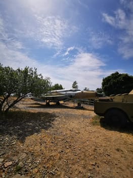 A military aircraft is positioned next to an old military vehicle in a rural area, surrounded by trees and under a bright sky, showcasing its historical significance and design.
