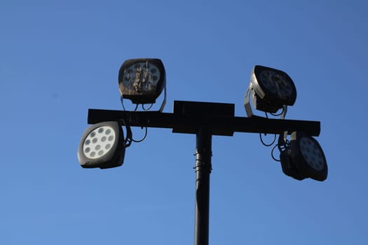 Four powerful stadium lights mounted on a pole shine brightly against a blue sky. These lights are typically used for evening sports events and activities.