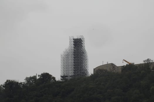 A tall building is enveloped in scaffolding, indicating ongoing renovations. The sky is overcast, and the structure is situated amidst a hilly landscape.