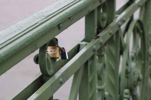 A small padlock attached to a green metal railing on a bridge reflects the tradition of lovers securing their commitments. Light rain adds a serene ambiance to the scene.
