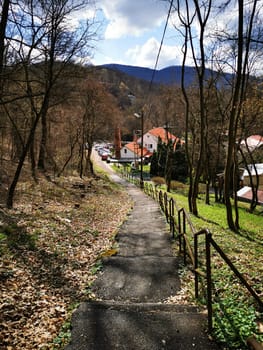 The path meanders down through bare trees, leading to a charming village nestled in the mountains, bathed in sunlight on a clear day.