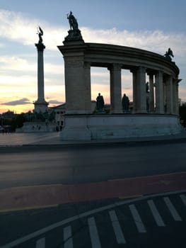 At sunset, a stunning colonnade and monument rise in a lively city square, showcasing intricate statues against a colorful sky, inviting visitors to explore its beauty.