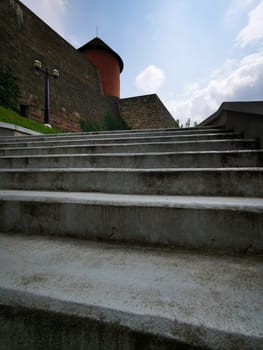 The concrete steps ascend toward an ancient fortress wall, surrounded by greenery, under a bright blue sky with fluffy clouds on a sunny afternoon.