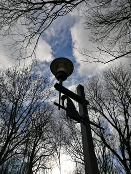 A tall lamp post is prominently positioned amidst leafless trees, surrounded by a dramatic cloudy sky as the sun sets in the background, illuminating the scene softly.