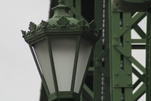 An intricately designed green street lamp stands on a bridge, showcasing decorative elements and ambient lighting, surrounded by industrial structures against a cloudy sky.