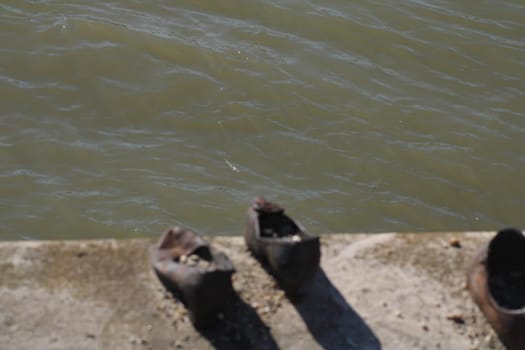 A pair of weathered shoes rests on the edge of a riverbank, partially submerged in murky water. The sunlight reflects off the river, enhancing the mood of abandonment.