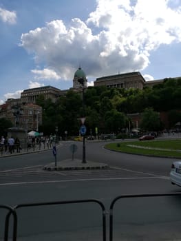 Visitors enjoy a leisurely day near Buda Castle, marveling at the architecture and natural surroundings under a clear blue sky with fluffy clouds.