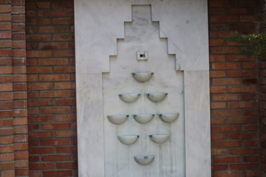 The wall fountain showcases a striking design with multiple bowls arranged in a geometric pattern. The structure is set against a rustic brick background, highlighting its elegance.