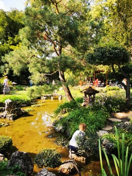 In a tranquil garden, a child bends down to explore a pond while lush greenery and stone structures create a peaceful atmosphere, inviting visitors to enjoy nature.