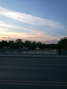 A tranquil scene unfolds at dusk, where a quiet bridge spans a shimmering lake, illuminated by the warm colors of the sunset against a clear sky.