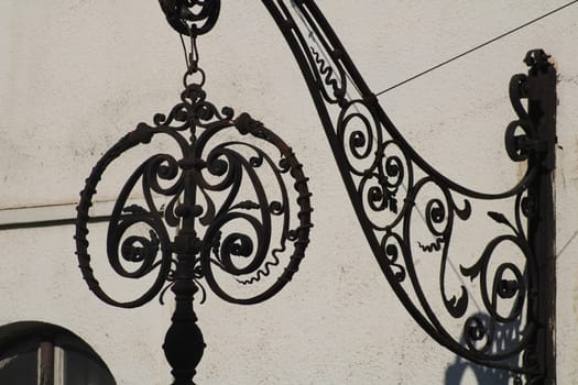 An elaborately crafted wrought iron lamp is suspended from a beautifully detailed bracket, casting shadows against a clean white wall in the daylight.