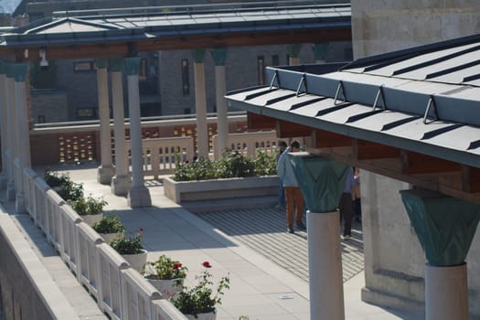 A serene courtyard features individuals strolling among colorful flowers and artistic architecture, showcasing an inviting outdoor space on a bright day.