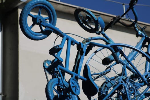 A collection of blue bicycles is creatively stacked against a pale wall, showcasing various sizes and shapes under a bright blue sky on a sunny day.