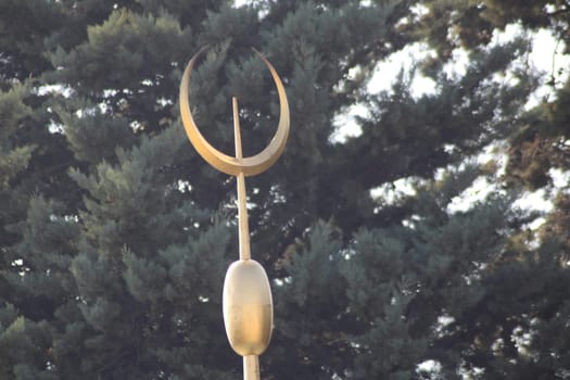 A striking golden crescent moon sculpture is perched high on a pole, framed by tall evergreen trees, capturing the warm glow of the setting sun in the background.