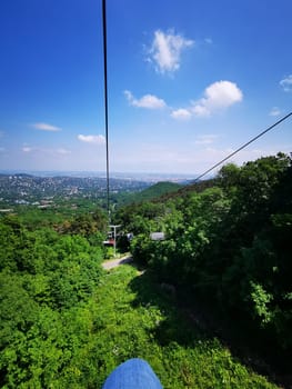 A cable car glides smoothly up the hillside, offering a breathtaking view of the city in the distance beneath a bright blue sky filled with fluffy clouds.