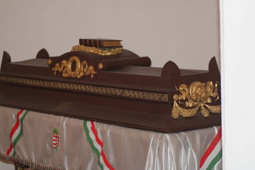 A beautifully crafted wooden coffin with gold accents rests in a quiet space, adorned with a decorative cloth featuring colorful ribbons, creating a respectful atmosphere.