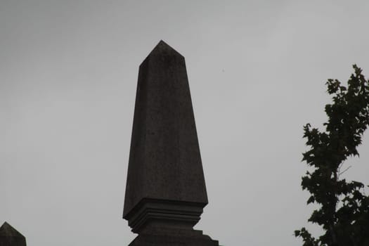 A tall obelisk rises prominently in a serene cemetery, silhouetted against a gray, overcast sky during the quiet moments of dusk.