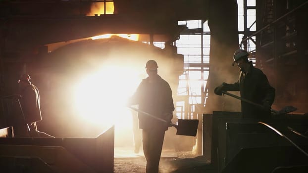 Silhouetted workers diligently laboring in a bustling steel mill, with molten metal glowing intensely in the background. Stock Clip