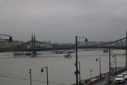 On a foggy evening in Budapest, boats glide along the Danube River beneath the Liberty Bridge, while the city’s skyline rises under a gray sky.