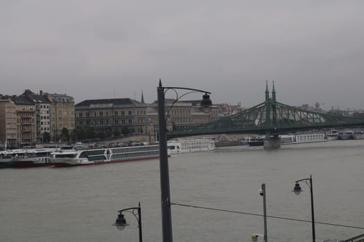 A cloudy afternoon in Budapest showcases the Danube River with a historic bridge and moored boats along the riverbank, reflecting the city's architectural beauty.