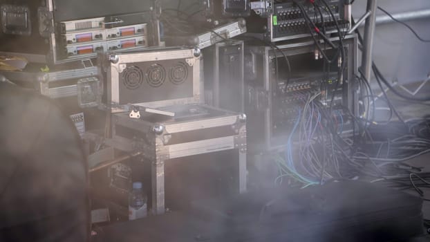 A detailed closeup view of professional audio and lighting equipment set up backstage featuring cables and fog. Clip