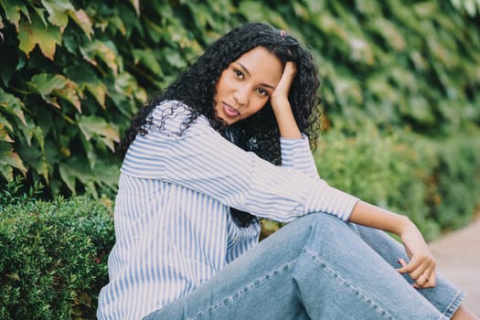 A young woman with flowing, curly hair exudes confidence and charm as she poses gracefully amidst vibrant greenery, embodying the ideals of modern beauty and a cheerful youthful spirit