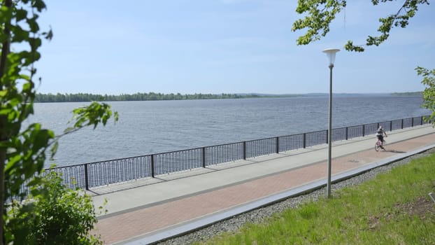 On this wonderful day, a cyclist is riding by the scenic waterfront, surrounded by lush greenery and bright blue skies. Clip