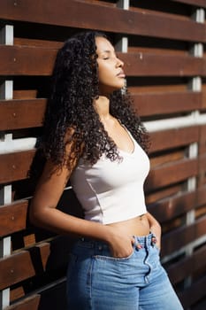 A young woman with beautiful curly hair delights in the warm golden sunlight as she leisurely leans against a rustic wooden fence, radiating a relaxed yet stylish and confident vibe