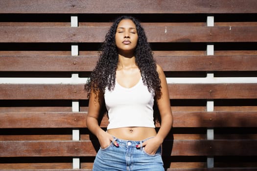 A vibrant young woman with curly hair stands confidently with her hands casually placed in her pockets, exuding a strong sense of strength and inner serenity against an earthy wooden backdrop