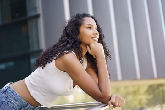 A young woman with curly hair gazes thoughtfully into the distance, leaning against a shiny railing in a vibrant outdoor setting filled with lush greenery and vivid colors, enjoying the moment