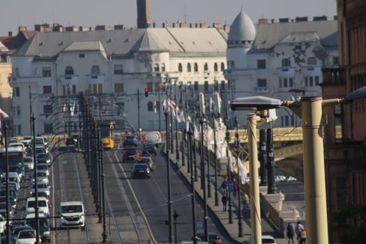 Cars traverse a bustling urban bridge lined with decorative flags. Historical buildings provide a stunning backdrop, showcasing the city's architecture in daylight.