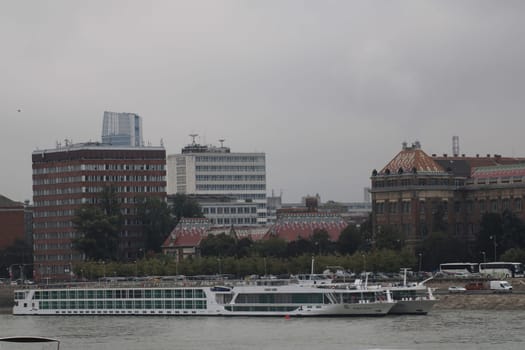 A river cruise ship sails along the Danube River in Budapest, surrounded by a mix of modern and historical buildings under overcast skies.
