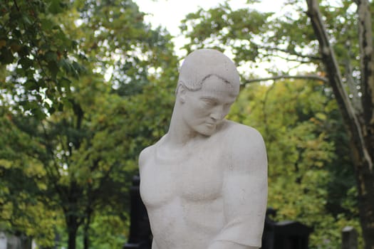 A detailed marble statue depicts a young man in a pensive pose, surrounded by vibrant green trees in a serene cemetery setting during the day.