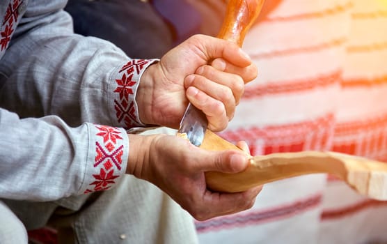 A skilled craftsman, clad in a white shirt adorned with red embroidery, meticulously carves a wooden spoon with a sharp knife. Wood shavings scatter around him, hinting at his dedication and the slow, meticulous process. The warm, natural light creates a sense of focus and tranquility as he works.