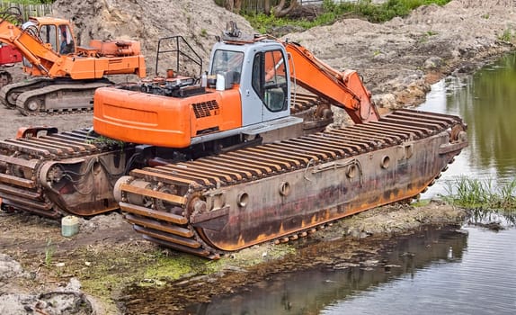 Two excavators, one parked and the other with its arm extended, sit in a dirt lot. Two workers are standing near the parked excavator, possibly making repairs.