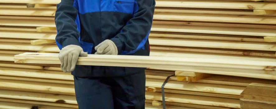 A worker carefully maneuvers a long wooden plank in a busy workshop, surrounded by neatly stacked lumber during daylight.