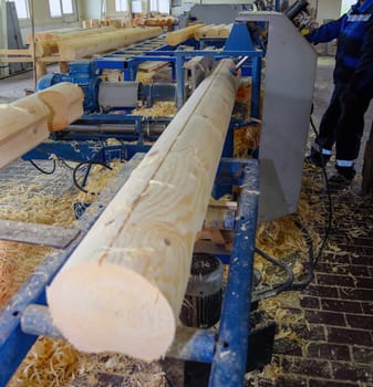 A worker stands at a wood processing machine, shaping logs from rough timber into smooth pieces in a busy workshop.