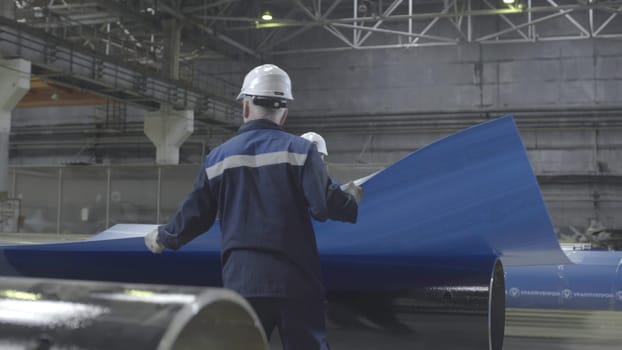 This video shows a large warehouse filled with metal pipes, featuring a skilled worker in a hard hat handling various pieces, including a blue pipe