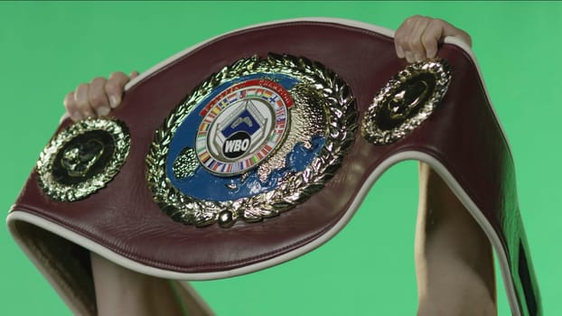 A person is confidently holding a boxing belt in front of a vibrant green screen backdrop, showcasing their achievement