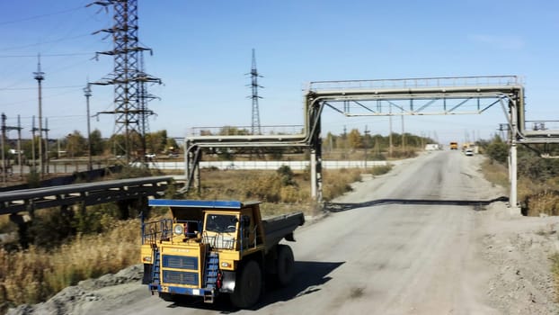 A vibrant blue and yellow dump truck is driving steadily on a dusty and unpaved road beneath a robust metal framework. Stock Clip