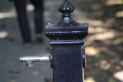 A decorative vintage water fountain stands alongside a shaded park pathway as pedestrians stroll by, enjoying the outdoor atmosphere in daylight.